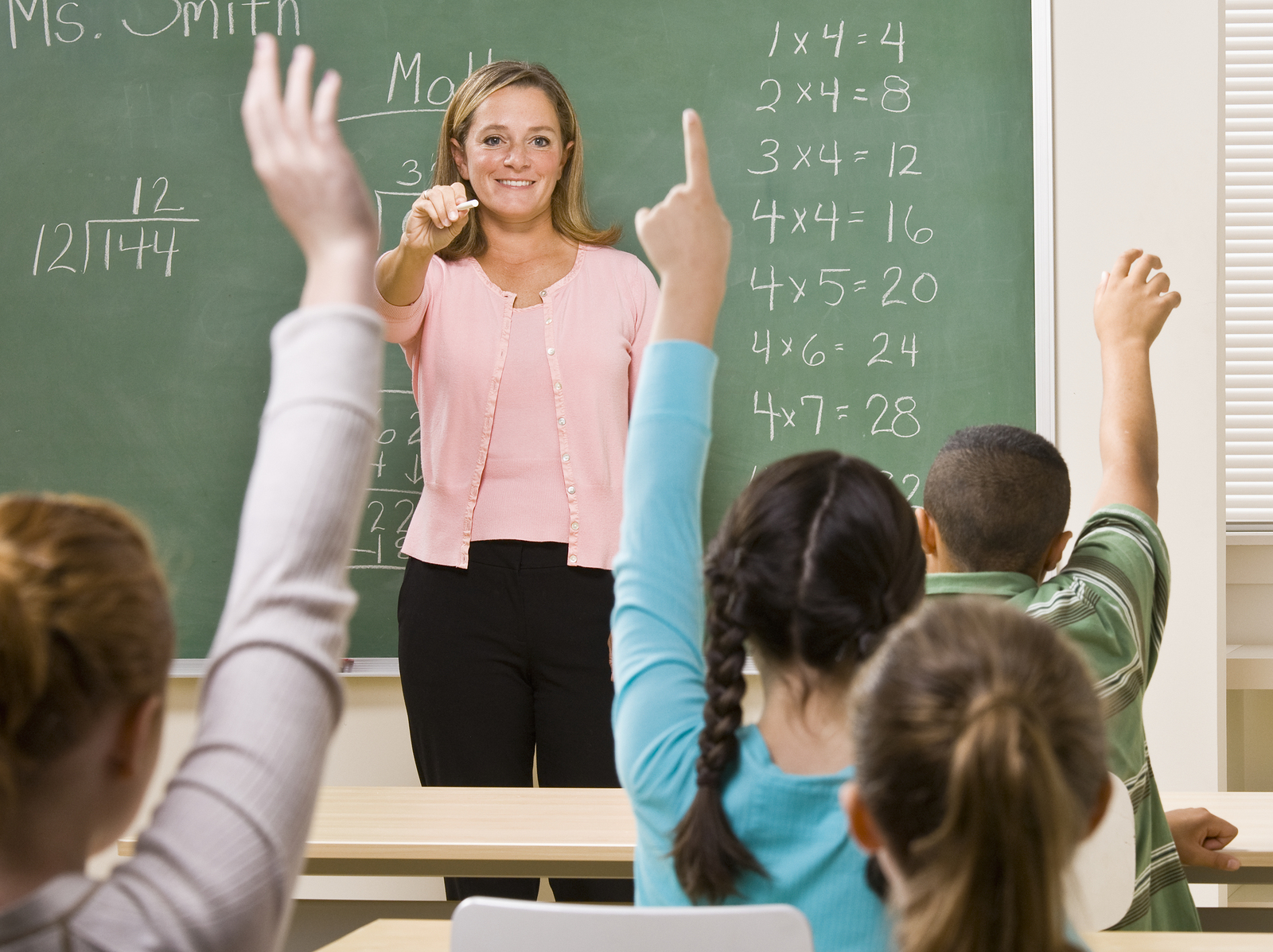  A teacher is standing in front of a blackboard with her students. She is smiling and looking at the students while holding a piece of chalk. Several students are raising their hands.